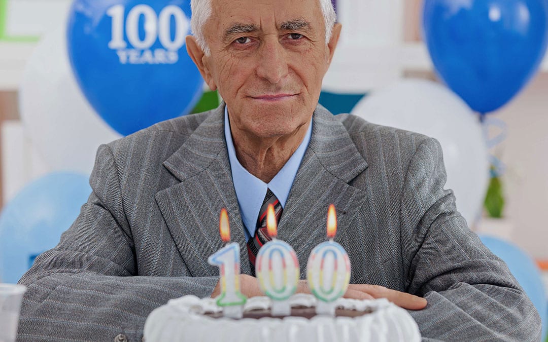 Older man sits by cake with candle that says 100